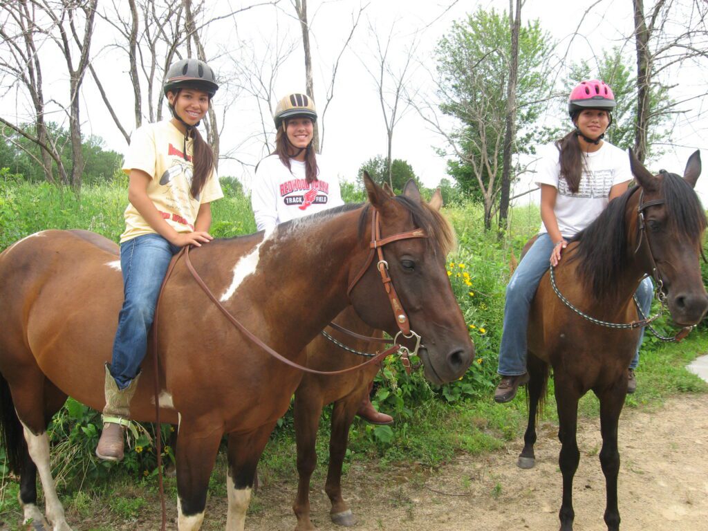 Three girls wearing helmets are sitting on two brown horses on a dirt path, surrounded by green grass and trees. They are smiling at the camera.