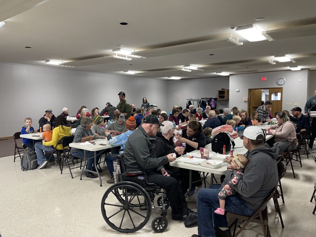 A large group of people, including children and adults, sit at tables in a community hall, eating and socializing. Some people are in wheelchairs and winter clothing is visible. The atmosphere is casual and friendly.