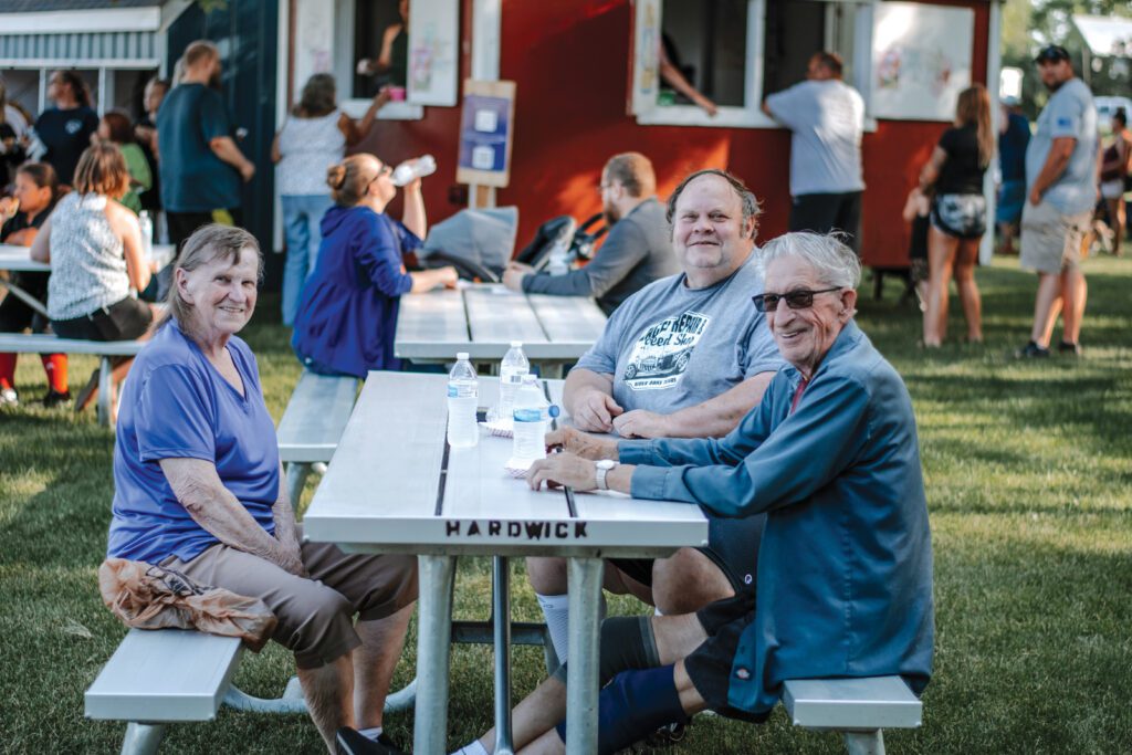 Three people sit at a white picnic table on grass, smiling at the camera. Other people are in the background near a red food stand. It is sunny, and water bottles are on the table.