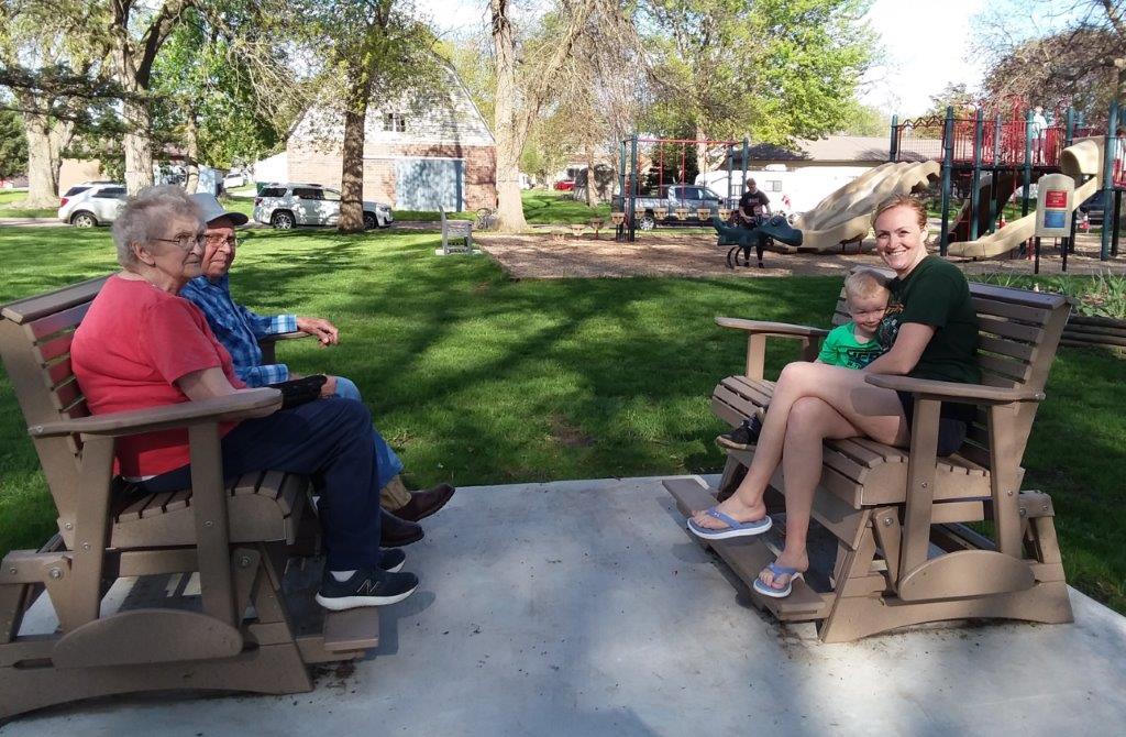 Two older adults sit on a glider bench facing a woman holding a small child on another bench in a park with trees, grass, and a playground in the background on a sunny day.