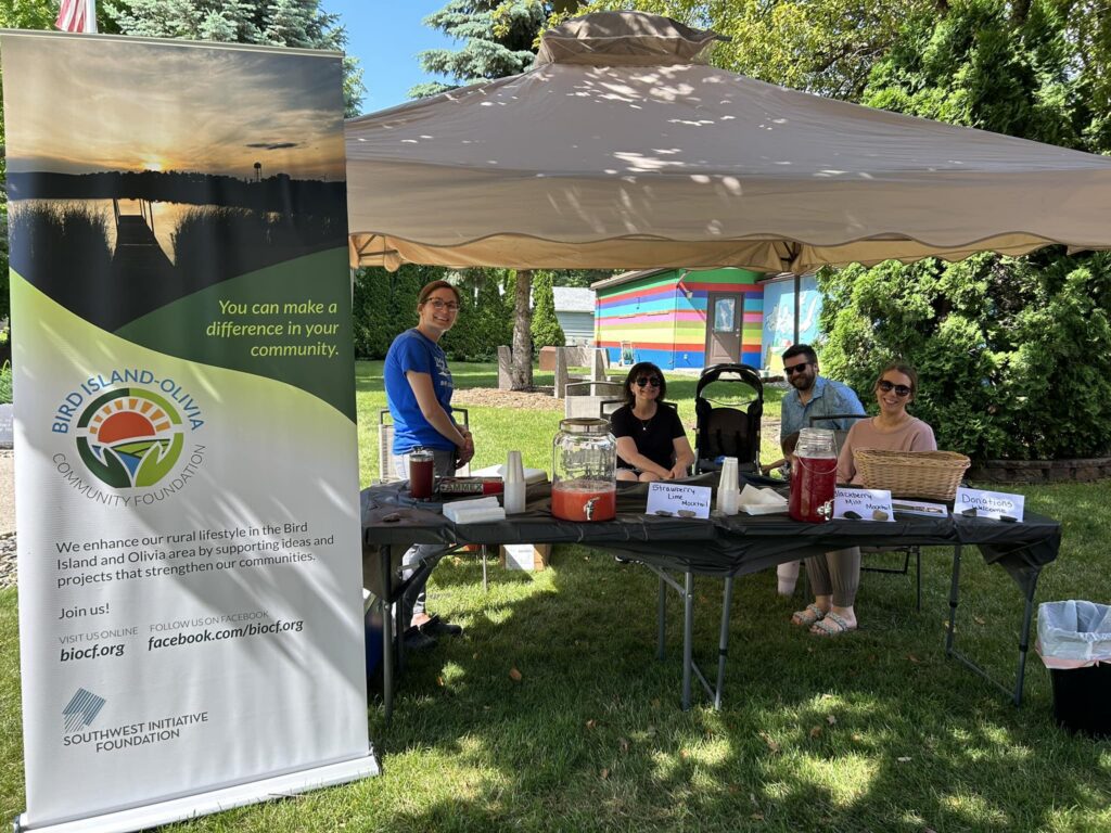 Four people sit and stand at a shaded outdoor booth with informational materials and drinks. A banner for the Bird Island-Olivia Community Foundation stands nearby. Trees and grass surround the area on a sunny day.