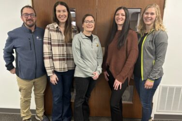Five people stand indoors in front of wooden double doors, smiling at the camera. They are dressed casually in sweaters, jackets, and jeans. The group appears to be in an office or workplace setting.