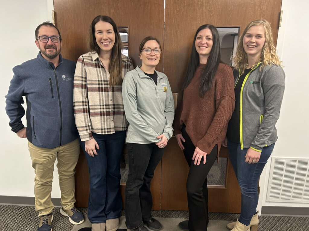 Five people stand indoors in front of wooden double doors, smiling at the camera. They are dressed casually in sweaters, jackets, and jeans. The group appears to be in an office or workplace setting.