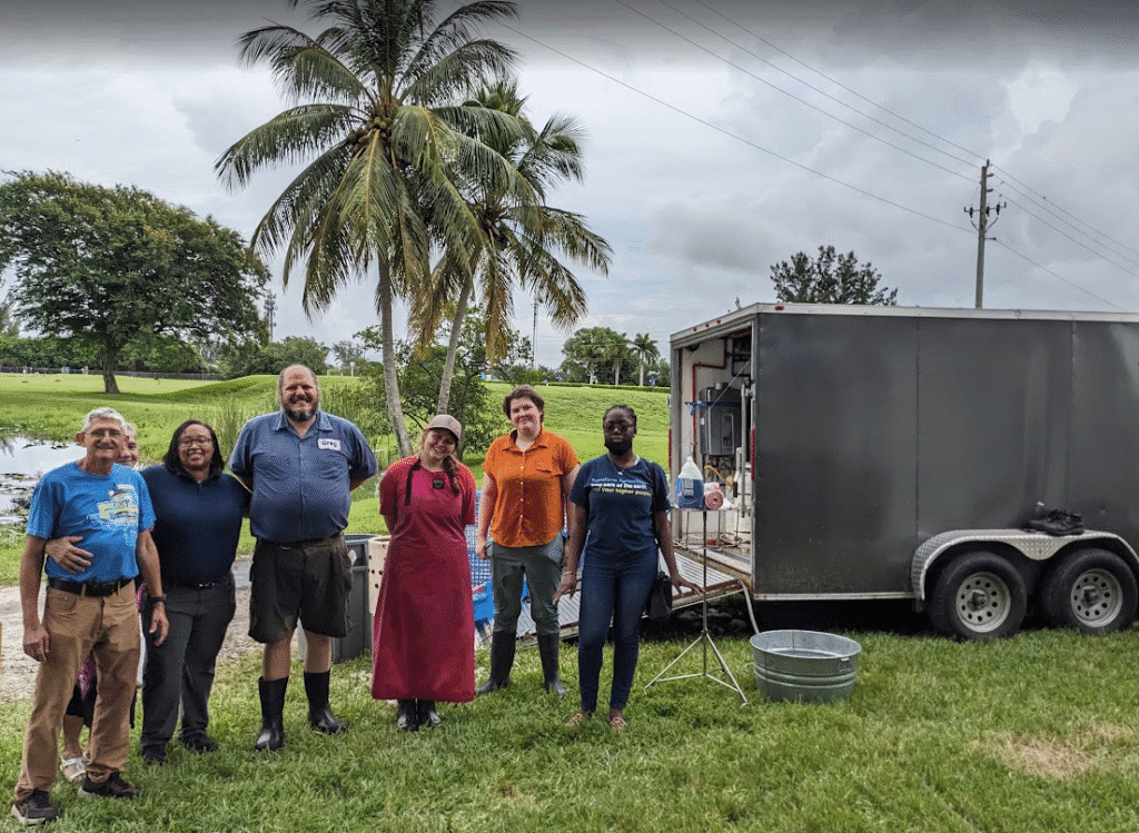 Six people stand together smiling on grass near a silver trailer and palm trees, with a pond and overcast sky in the background. Some wear boots and aprons, suggesting an outdoor or agricultural activity.