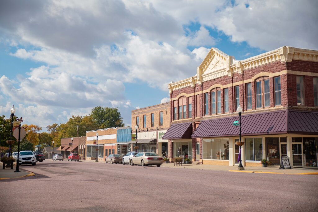 A small town main street with historic brick buildings, cars parked along the road, shops with large windows, and a partly cloudy sky overhead. Trees with autumn leaves are visible in the background.