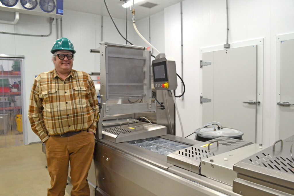 A man wearing a green hard hat, safety glasses, and plaid shirt stands in a clean, industrial food processing facility next to a metal packaging machine and control panel.