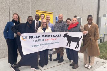 A group of ten people stand outside a building, smiling and holding a large banner that reads, “FRESH GOAT SALE 8 am-4:30 pm on Fridays! First come, first served unless pre-ordered,” with a goat silhouette on it.