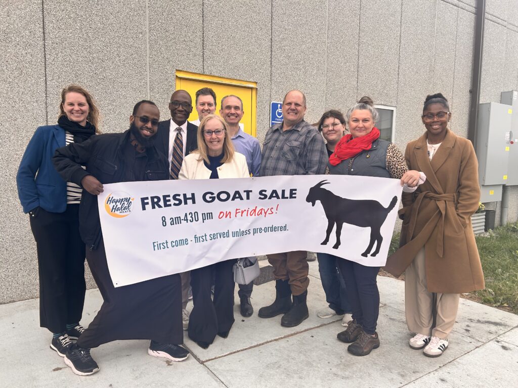 Ten people stand outside a building, smiling and holding a banner that reads “Fresh Goat Sale 8 am-4:30 pm on Fridays! First come, first served unless pre-ordered.” A goat silhouette is printed on the banner.