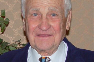 An elderly man with white hair, wearing a dark blue blazer, white shirt, and a colorful patterned tie, smiles at the camera. A leafy plant and textured wall are visible in the background.
