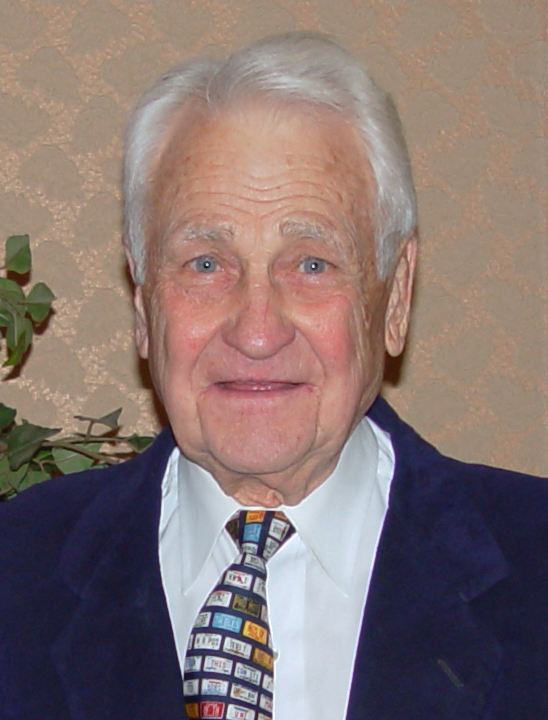An elderly man with white hair, wearing a dark blue blazer, white shirt, and a colorful patterned tie, smiles at the camera. A leafy plant and textured wall are visible in the background.