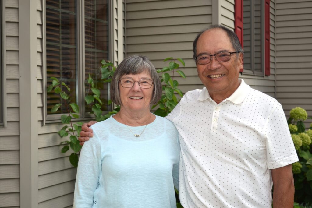 An older woman and man stand smiling together outside a house with beige siding and greenery. The man has his arm around the woman; both wear glasses and casual clothing.