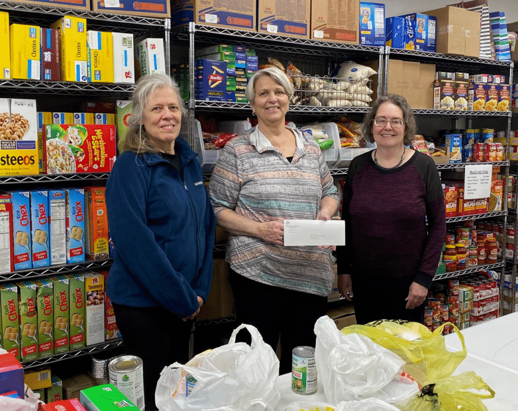 Three women stand and smile in front of shelves filled with boxed and canned food, utilizing nonprofit resources. One holds an envelope, with plastic bags and canned goods on the table, suggesting a food pantry or donation setting.