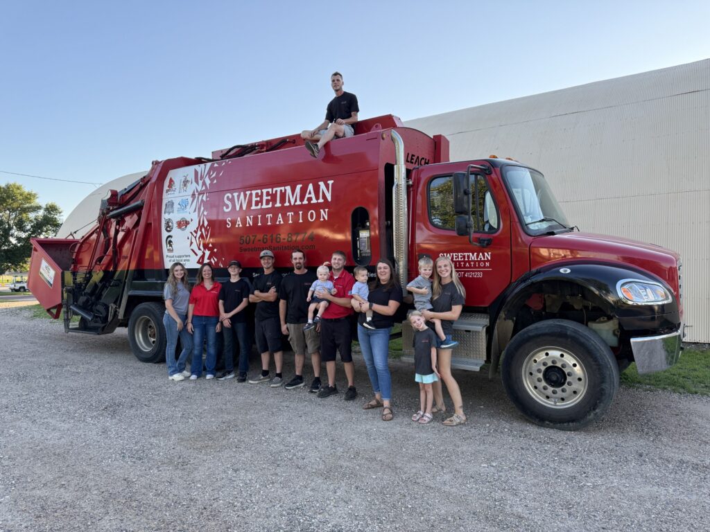 A group of people, including adults and children, stand smiling in front of a red Sweetman Sanitation garbage truck. One person sits on top of the truck. The scene is outdoors on a gravel surface.