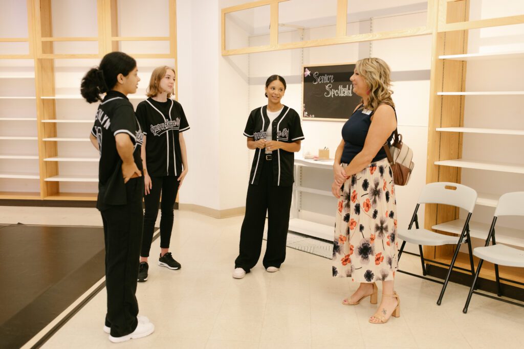 Four women stand in conversation in a room with empty shelves. Three wear matching black shirts and pants, while one, dressed in a floral skirt and sleeveless top, faces them, smiling. Folded chairs are nearby.