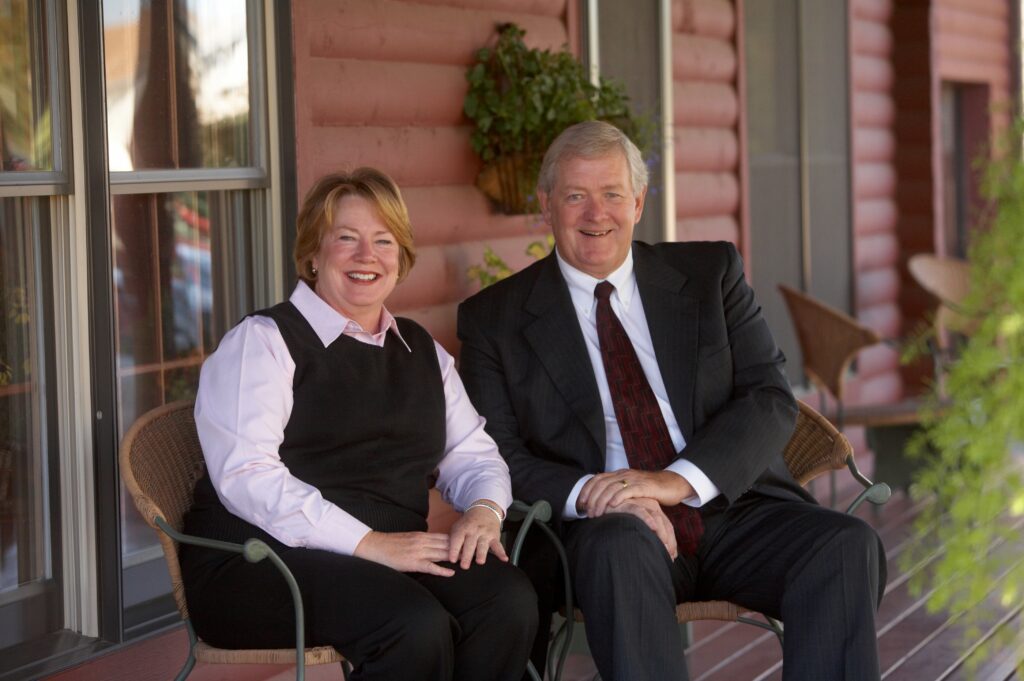A smiling man and woman in business attire sit side by side on chairs, outside a red log building with windows and plants in the background.