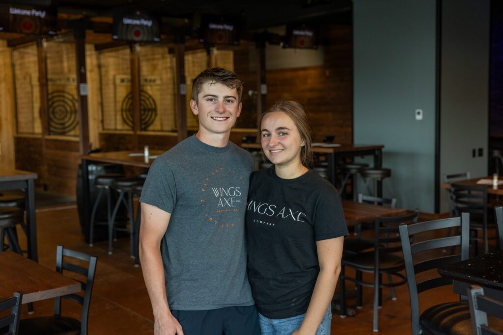 Two young adults stand smiling together inside an axe throwing venue, wearing matching WINGS AXE t-shirts. Empty tables and wooden axe targets are visible in the background.