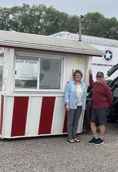 Man and woman happily standing against a small white and red striped booth in a parking lot near some equipment.
