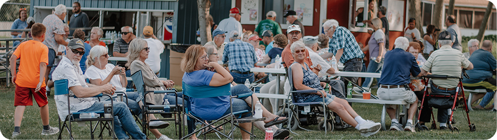 A group of older adults gather outdoors, sitting on folding chairs and benches, socializing and enjoying a community event on a grassy area near food stands.