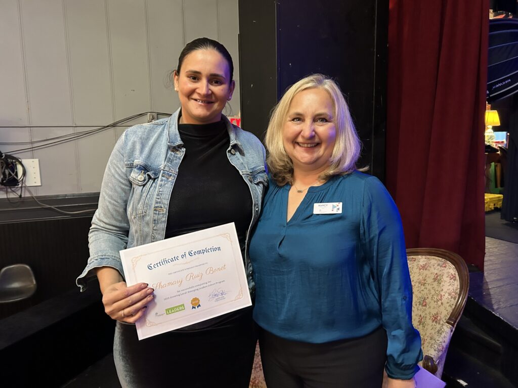 Two women smile at the camera; one holds a certificate of completion. They stand indoors near a vintage chair, with a red curtain and dark walls in the background.