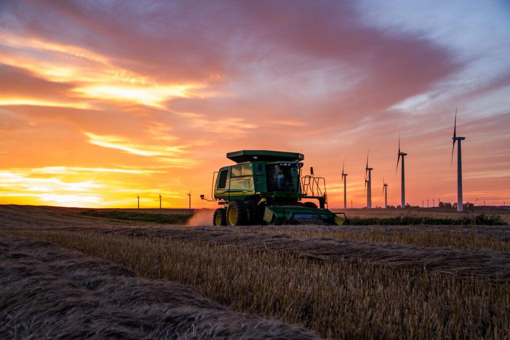 A green combine harvester works in a field at sunset, with orange and pink clouds in the sky and wind turbines in the background.