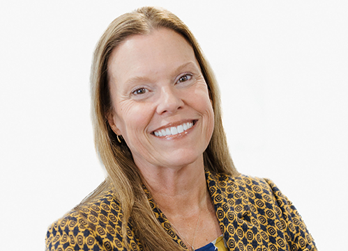 Stacy has straight light brown hair and is wearing a patterned blazer over a blue top, posed in front of a plain white background smiling.