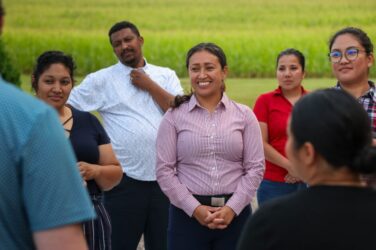 A group of people stand outdoors in front of green fields, smiling and talking together. One woman in a striped shirt stands at the center, appearing to lead or address the group.