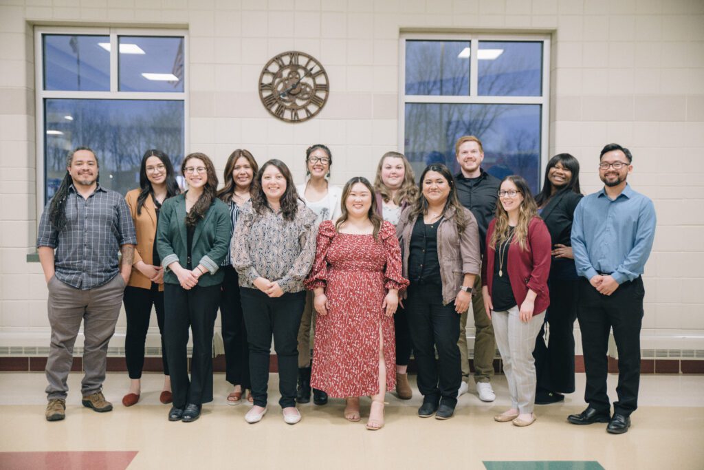 A group of fourteen adults, dressed in business casual attire, stand smiling in two rows inside a building with large windows and a clock on the wall behind them.