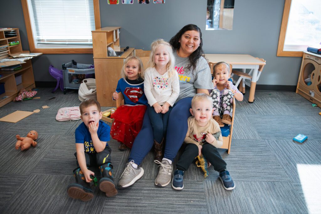 A smiling woman sits on the floor of a classroom with five young children around her. The children appear happy and playful, with toys and classroom furniture visible in the background.