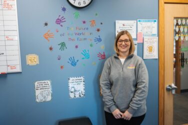 A woman with glasses and a gray sweater stands smiling in a classroom by a wall with colorful handprints and a sign that says, The Future of the World is in This Room. Various posters and a clock are also visible.
