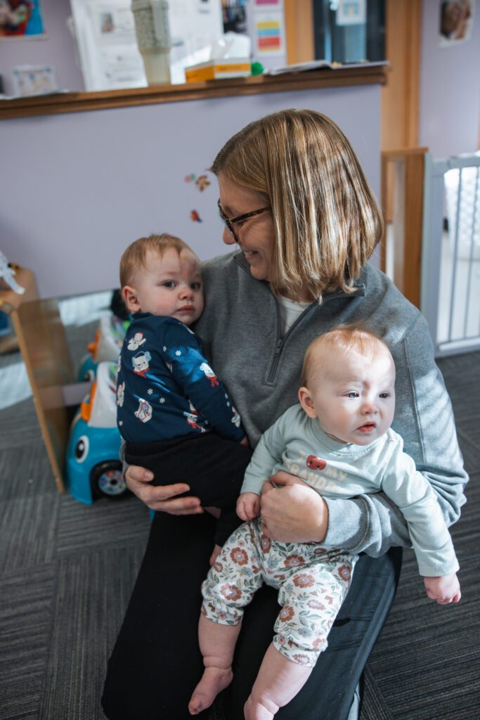 A woman with glasses and shoulder-length hair sits on the floor, smiling and holding two babies on her lap. Both babies are wearing printed outfits, and toys and artwork are visible in the background.