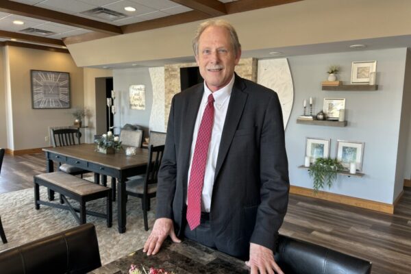 A man in a dark suit and red tie stands smiling in a modern, warmly decorated dining room with wooden beams, a dining table, wall art, and shelves with plants and decor.