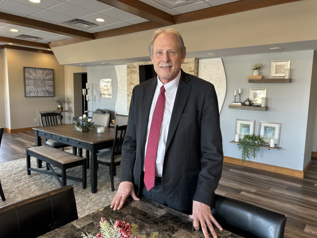 A man in a dark suit and red tie stands smiling in a modern, warmly decorated dining room with wooden beams, a dining table, wall art, and shelves with plants and decor.