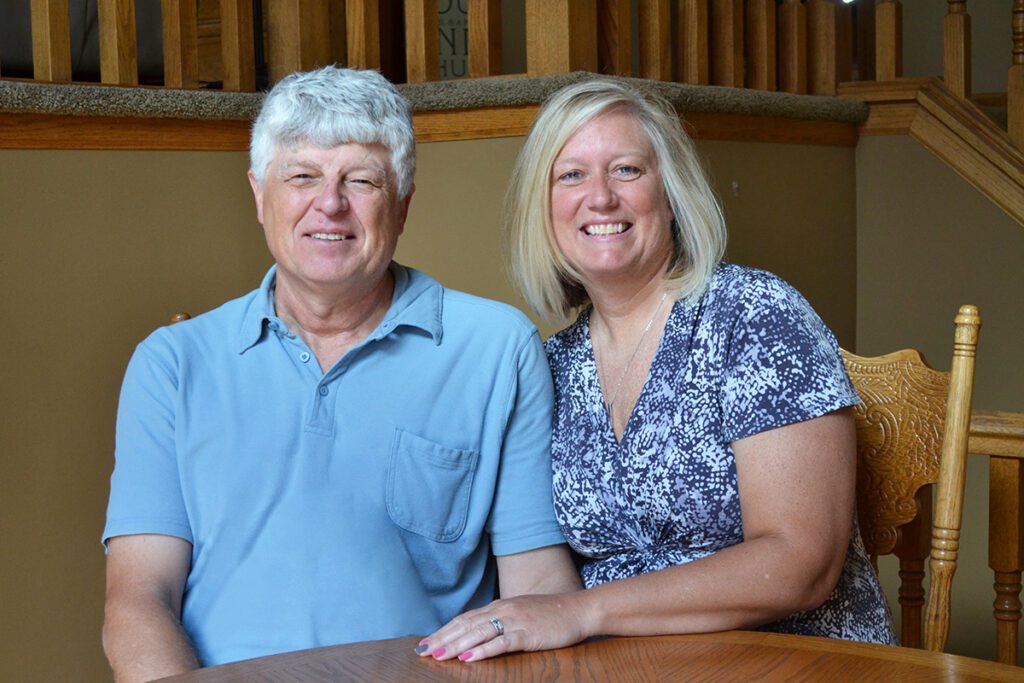 A smiling man and woman sit together at a wooden table in a warmly lit home, with stairs and wooden railings in the background. The man wears a light blue polo shirt, and the woman wears a patterned dress.