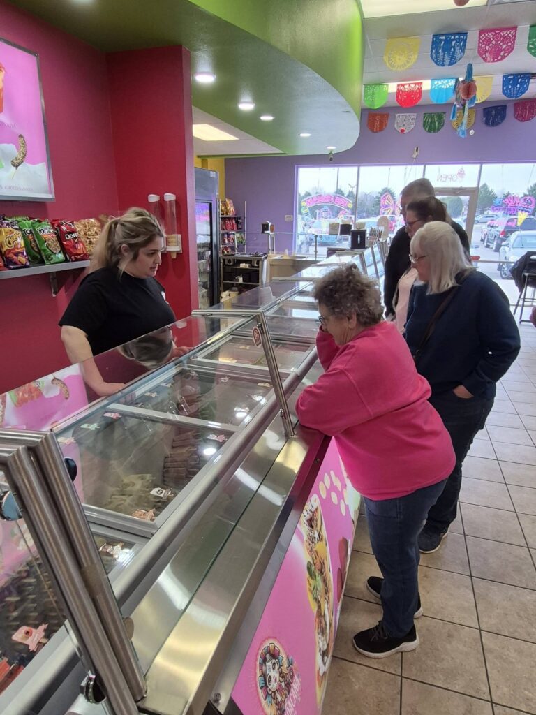 Three people stand at an ice cream shop counter, looking at the options while a staff member behind the glass display waits to take their order. Colorful decorations hang from the ceiling.