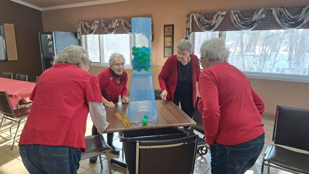 Four elderly women, all wearing red tops, are gathered around a table playing a game with a tall plastic tower containing green and blue balls and yellow sticks. 