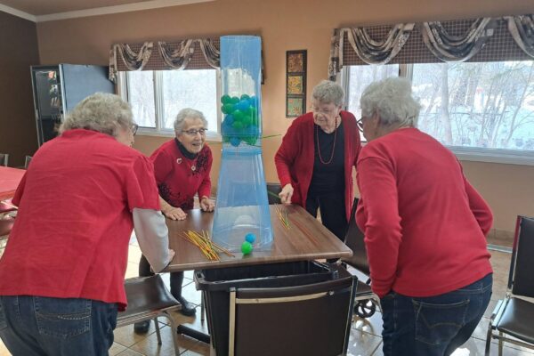 Four elderly women, all wearing red tops, are gathered around a table playing a game with a tall plastic tower containing green and blue balls and yellow sticks.