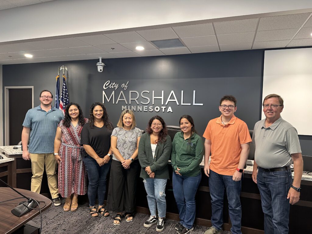 Eight people stand side by side, smiling in front of a wall with the words “City of Marshall Minnesota.” An American flag is in the background, and they appear to be in a meeting or conference room.
