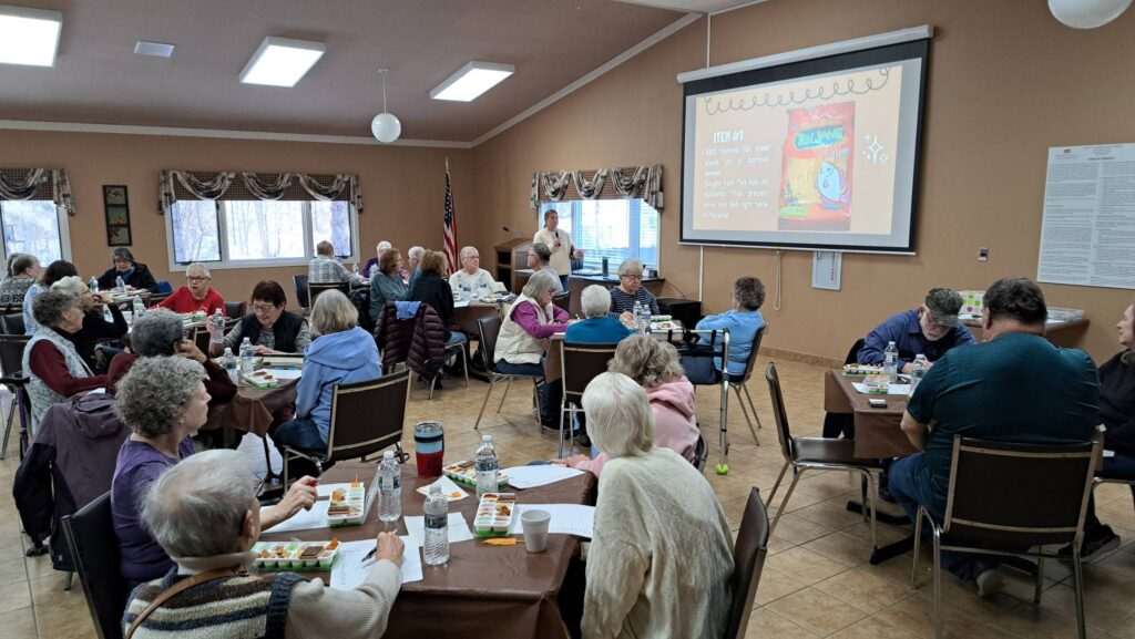 A group of seniors sit at tables in a community room eating lunch while a woman at the front gives a presentation projected on a large screen. 