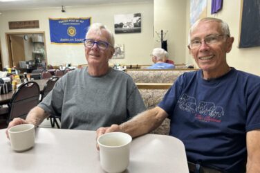 Two older men sit at a table in a diner, each holding a white coffee mug and smiling at the camera. The diner has simple decor, and an American Legion banner hangs on the wall in the background.