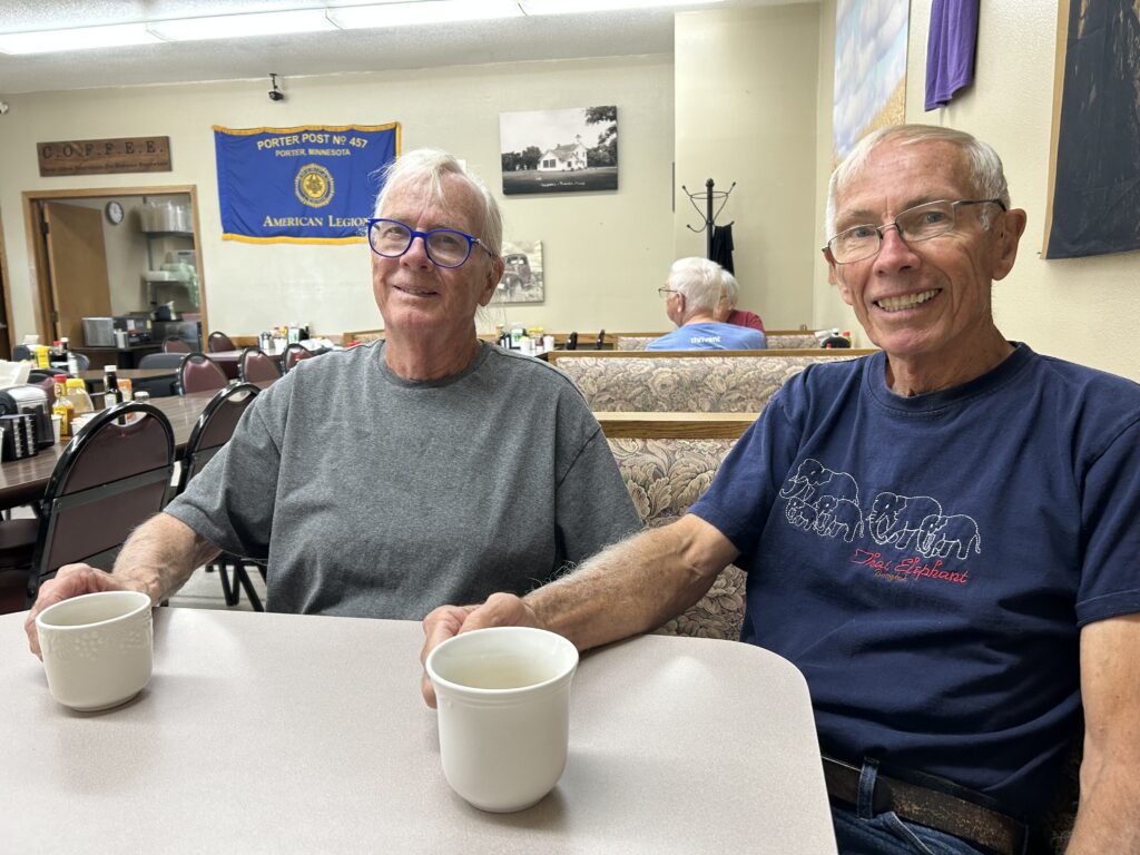 Two older men sit at a table in a diner, each holding a white coffee mug and smiling at the camera. The diner has simple decor, and an American Legion banner hangs on the wall in the background.