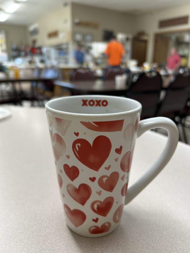 A white mug with red heart designs and XOXO printed inside sits on a table in a cafe, with blurred people and tables in the background.
