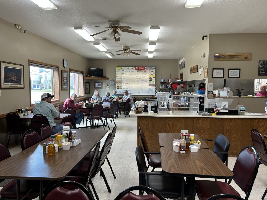 A small diner with several tables and chairs, a counter with condiments and a coffee machine, people dining, and a large menu on the back wall. The atmosphere is casual and cozy with natural light from windows.