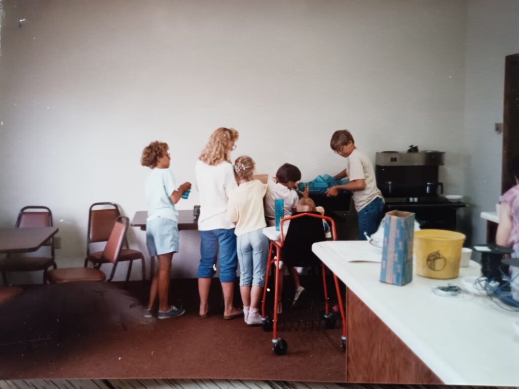 A group of children stand together by a counter in a room with tables and chairs. One child uses a red walker, and others appear to be helping or talking. A yellow bucket sits on the counter in the foreground.