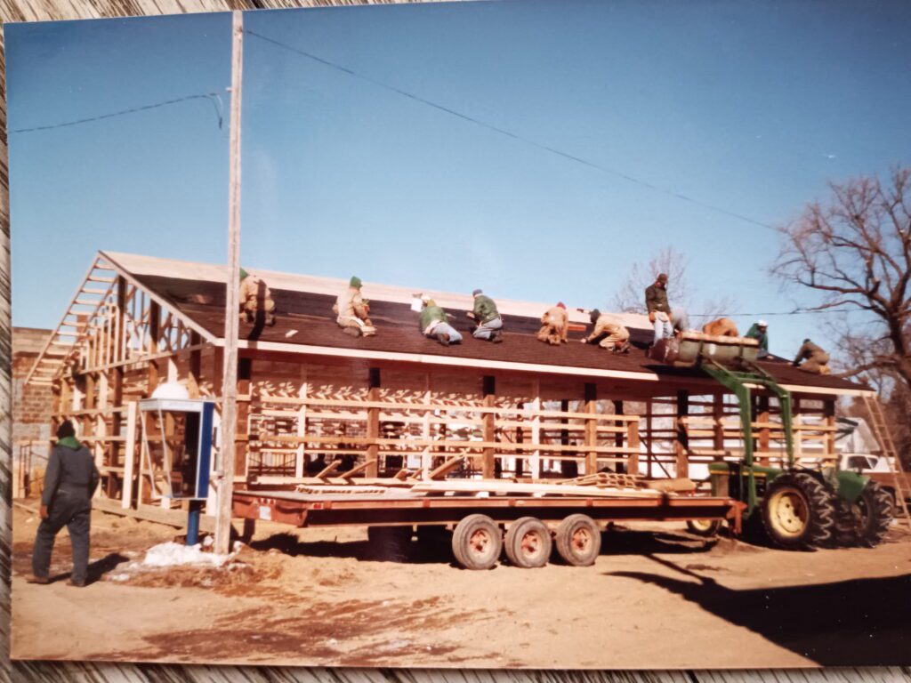 Several workers are installing shingles and working on the roof of a large wooden building under construction. A tractor with a trailer is parked in front, and another person stands on the ground nearby. The sky is clear and blue.
