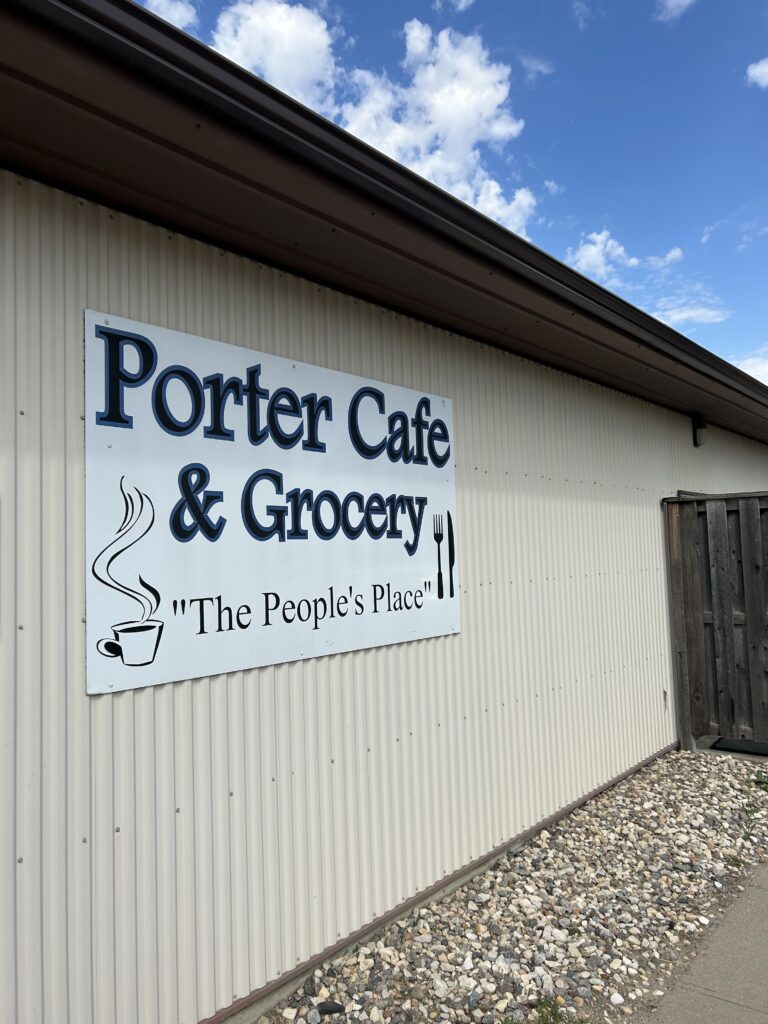 A sign on a corrugated building reads Porter Café & Grocery The Peoples Place with images of a steaming coffee cup, a fork, and a knife. The sky above is partly cloudy.