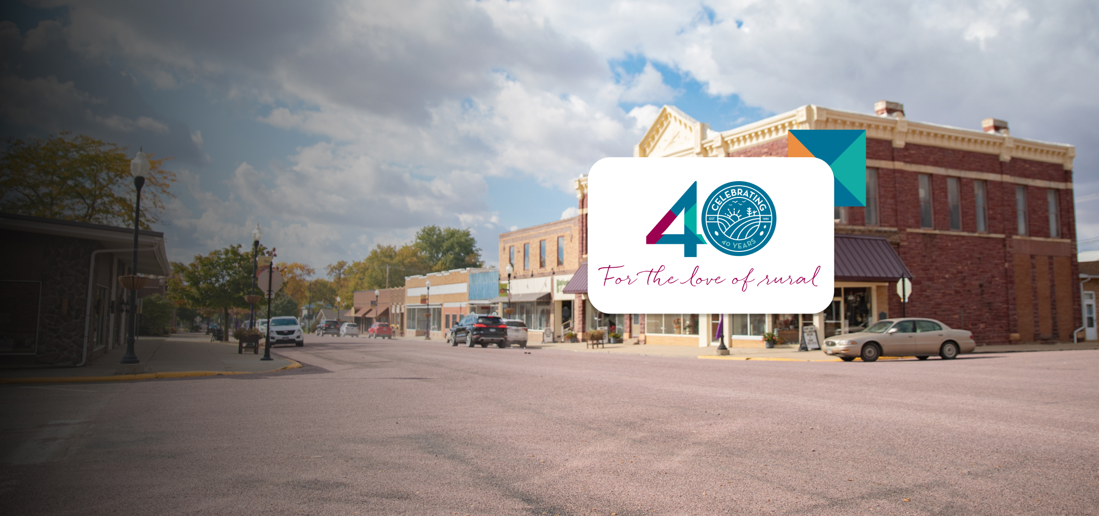 A small town street with historic brick buildings under a partly cloudy sky. Overlaid is a logo reading 40 For the love of rural with a blue seal and colorful accent.
