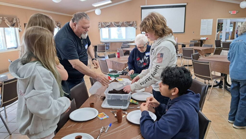 A group of people of various ages gather around a table in a community room, working together on a gardening project with soil, seeds, and small pots. Plates and craft supplies are also on the table.