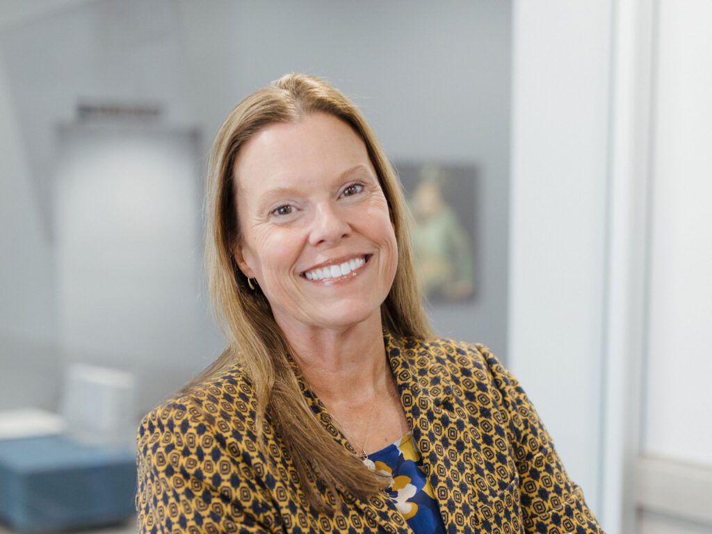 A woman with long light brown hair smiles at the camera. She is wearing a patterned blazer over a blue top, and the background is a softly blurred indoor office or professional setting.