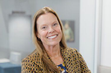 Stacy has long light brown hair and smiles at the camera. She is wearing a patterned blazer over a blue top, and the background is a softly blurred office.