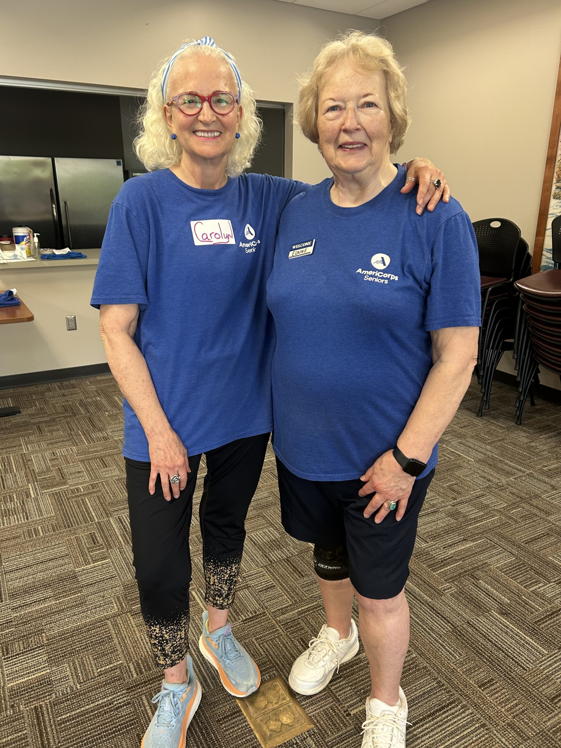 Two smiling older women wearing matching blue shirts and name tags stand arm-in-arm in a room with brown carpet, chairs stacked in the background, and a countertop with snacks.